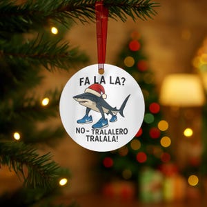 May include: A round, white Christmas ornament with a shark wearing a Santa hat and sneakers. The text "FA LA LA?" and "NO - TRALALERO TRALALA!" are printed on the ornament. The ornament hangs from a red ribbon.