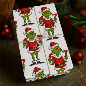 Pode incluir: Presente embrulhado com um padrão repetido de um personagem de desenho animado verde em um traje de Papai Noel segurando os números 6 e 7. O presente é amarrado com barbante e repousa sobre uma superfície de madeira. Enfeites de Natal vermelhos ao fundo.