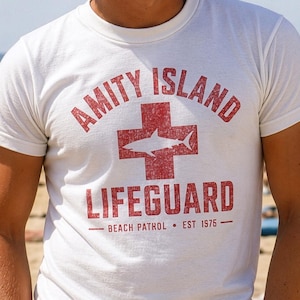Könnte beinhalten: Weißes T-Shirt mit einem roten, verblassten Aufdruck. Der Aufdruck lautet "Amity Island Lifeguard" mit einem Hai in einem Kreuz, "Beach Patrol" und "Est 1976". Das Shirt hat kurze Ärmel.