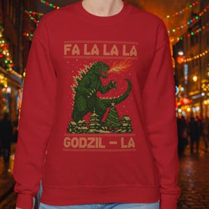 Può includere: Felpa rossa con un design pixelato di Godzilla verde che sputa fuoco. Il testo "FA LA LA LA" e "GODZIL - LA" è visualizzato in un carattere festivo. Un albero di Natale e degli edifici sono presenti nel design.