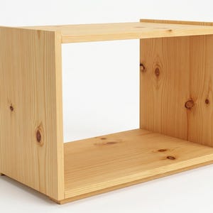 May include: A rectangular open-front storage unit constructed from light-coloured natural pine wood. The piece features visible wood grain patterns and several dark circular knots throughout the side and base panels, with a simple box-like frame.