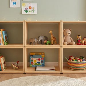 May include: A light-coloured wooden modular shelving unit with six open compartments. The shelves are filled with various children's books, a spinning top, animal figurines, a teddy bear, nesting dolls, a colourful woven basket, and a game box.