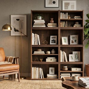 May include: A tall, dark wood double bookcase with multiple shelves filled with books, framed pictures, and decorative objects. The unit stands next to a tan leather armchair, a brass floor lamp, and a potted green plant on a woven rug.