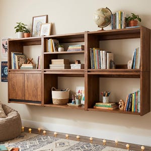 May include: A modular wall-mounted wooden shelving unit consisting of three connected sections with open shelves and a lower cabinet with two doors. The unit holds various books, a globe, small potted plants, a woven basket, and art supplies.