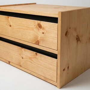 May include: A small wooden storage unit featuring two drawers with visible natural wood grain and knots. The drawer fronts are light brown, while the recessed horizontal gaps between the drawers and the top frame are finished in a solid matte black.
