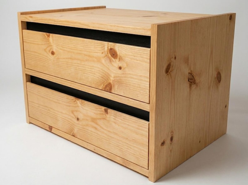 May include: A small wooden storage unit featuring two drawers with visible natural wood grain and knots. The drawer fronts are light brown, while the recessed horizontal gaps between the drawers and the top frame are finished in a solid matte black.