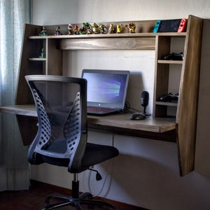 Op de afbeelding: Een zwarte mesh bureaustoel met een houten bureau dat aan de muur is bevestigd. Het bureau heeft een laptop, een microfoon en een muis erop. Boven het bureau bevinden zich planken met videogamefiguren en controllers.