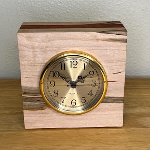 May include: A wooden quartz clock with a gold-coloured face and hands. The clock is set in a light-coloured, rectangular wooden block with a gold-coloured bezel. The wood grain is visible on the clock's face and the block's sides.