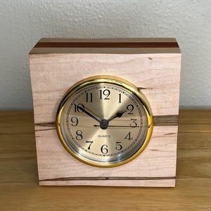 May include: A square wooden clock with a gold-coloured clock face and a light-coloured wood grain. The clock face has black numbers and hands. The clock is made of light-coloured wood with a darker wood accent.