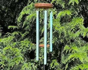 May include: A teal blue wooden wind chime with a square wooden base hanging from a tree branch. The wind chime has four tubes and a wooden top.