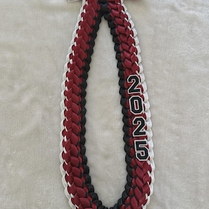 May include: A braided graduation lei in maroon, black, and white, with the year "2025" in black and white. The lei is tied with a maroon, black, and white ribbon bow.