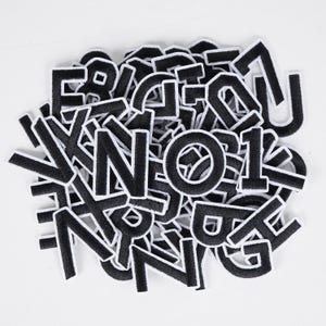 May include: A collection of black embroidered letters and numbers with white borders, scattered on a white surface. The letters and numbers are in a bold, sans-serif font, suitable for patches or embellishments.