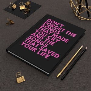 Op de afbeelding: Een zwart notitieboekje met de tekst "Vergeet de liedjes niet die je aan het huilen hebben gemaakt en de liedjes die je leven hebben gered" in roze letters.