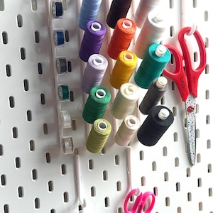 May include: A white pegboard with a white thread organiser holding spools of thread in various colours, including red, orange, yellow, green, blue, purple, and white. Two pairs of scissors are hanging on the pegboard, one red and one pink.