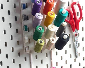 Peut inclure: Un panneau perforé blanc avec un organisateur de fil blanc contenant des bobines de fil de différentes couleurs, notamment rouge, orange, jaune, vert, bleu, violet et blanc. Deux paires de ciseaux sont accrochées au panneau perforé, une rouge et une rose.