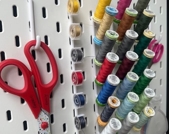 Peut inclure: Un panneau perforé blanc avec une variété de bobines de fil de couleurs organisées sur des crochets. Les bobines sont étiquetées avec la marque "Gutermann". Une paire de ciseaux rouges à pois est accrochée à un crochet.