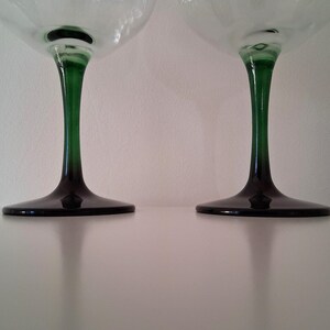 May include: Two stemmed glasses with clear bowls and dark green stems and bases. The stems transition from green to black at the base. The glasses are set against a white background.