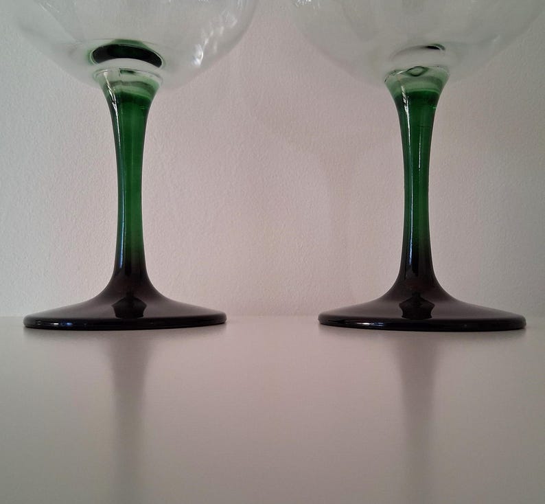 May include: Two stemmed glasses with clear bowls and dark green stems and bases. The stems transition from green to black at the base. The glasses are set against a white background.