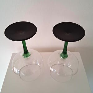 May include: Two clear wine glasses with a black disc base and a green stem. The glasses are upside down, resting on a white surface. The stems are a vibrant green color, contrasting with the clear bowl of the glass.