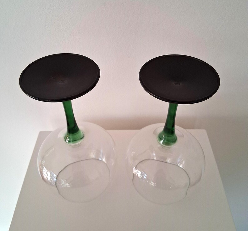 May include: Two clear wine glasses with a black disc base and a green stem. The glasses are upside down, resting on a white surface. The stems are a vibrant green color, contrasting with the clear bowl of the glass.