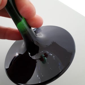 May include: A close-up of a wine glass with a dark, glossy bowl and a long, green-tinted stem. The glass is held by a hand, showcasing the elegant design and reflective surface. The bowl is a deep, dark color.