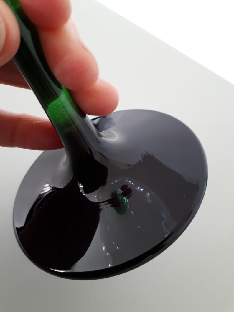 May include: A close-up of a wine glass with a dark, glossy bowl and a long, green-tinted stem. The glass is held by a hand, showcasing the elegant design and reflective surface. The bowl is a deep, dark color.