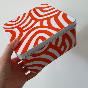 Könnte beinhalten: Eine quadratische Blechdose in Weiß und Orange mit einem geometrischen Muster. Die Dose ist aus Metall und hat einen Klappdeckel.