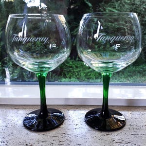 May include: Two clear gin glasses with green stems and black bases. The glasses have a rounded bowl shape and the word "Tanqueray" is printed in white script on the front of each glass. The glasses are sitting on a light-colored surface.