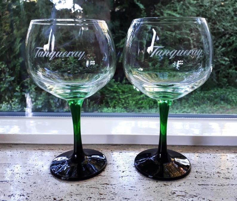 May include: Two clear gin glasses with green stems and black bases. The glasses have a rounded bowl shape and the word "Tanqueray" is printed in white script on the front of each glass. The glasses are sitting on a light-colored surface.