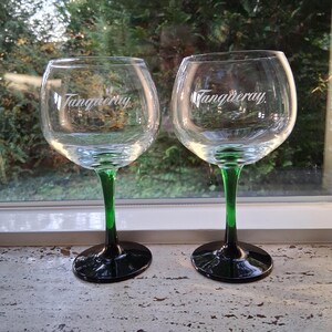 May include: Two clear gin glasses with the word "Tanqueray" in white script. The stems are green, transitioning to black at the base. The glasses are positioned in front of a window with a blurred green background.