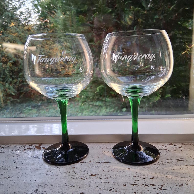May include: Two clear gin glasses with the word "Tanqueray" in white script. The stems are green, transitioning to black at the base. The glasses are positioned in front of a window with a blurred green background.