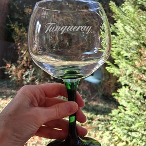 May include: A clear, balloon-shaped glass with a green stem and base, featuring the etched word "Tanqueray." The glass is held up against a blurred green background, suggesting an outdoor setting. The glass is designed for cocktails.