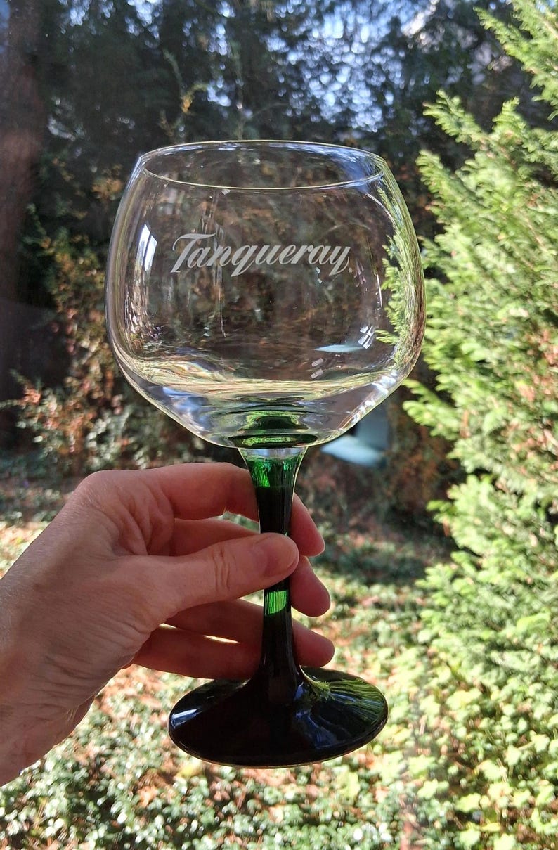 May include: A clear, balloon-shaped glass with a green stem and base, featuring the etched word "Tanqueray." The glass is held up against a blurred green background, suggesting an outdoor setting. The glass is designed for cocktails.