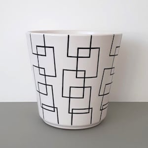 Könnte beinhalten: Weißer Keramiktopf mit geometrischem Schwarz-Weiß-Muster. Der Topf hat eine breite Basis und eine leicht konische Oberseite.