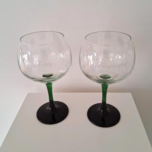 May include: Two clear, stemmed gin glasses with green stems and black bases. The glasses have a rounded bowl shape and the word "Tanqueray" is etched on the side of each glass. The glasses are sitting on a white surface.