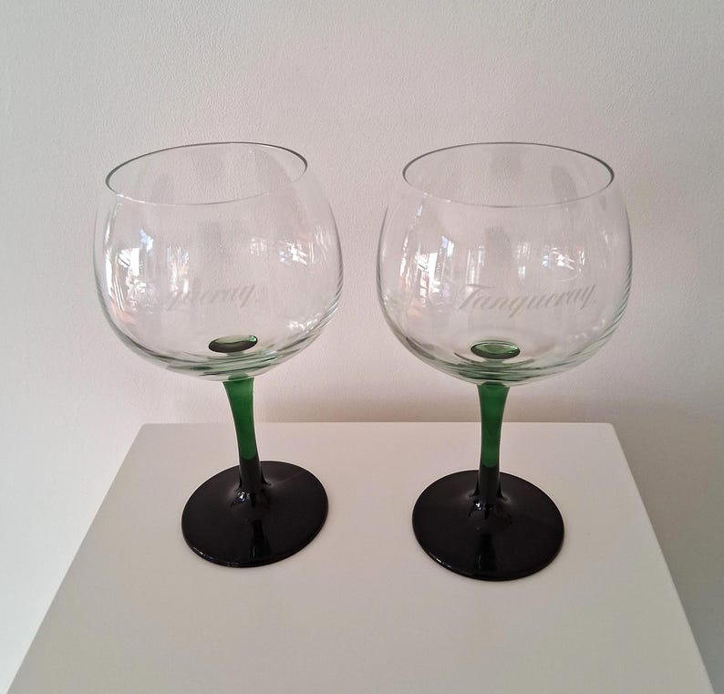 May include: Two clear, stemmed gin glasses with green stems and black bases. The glasses have a rounded bowl shape and the word "Tanqueray" is etched on the side of each glass. The glasses are sitting on a white surface.
