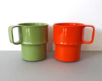 May include: Two brightly colored mugs. One is a vibrant green, the other a bold orange. Both have a cylindrical shape with a handle and a distinctive stacked design around the middle. The mugs are set against a plain white background.
