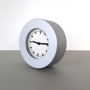 Puede incluir: Un reloj analógico redondo de color plateado con una esfera blanca y manecillas y marcas negras. El reloj tiene un diseño minimalista y se encuentra sobre una superficie gris. La esfera del reloj muestra la hora.