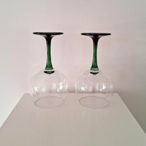 May include: Two clear wine glasses with a green stem and dark rim are displayed upside down. The glasses are on a white surface against a white background. The stem is a vibrant green color, and the rim is a dark, almost black color.