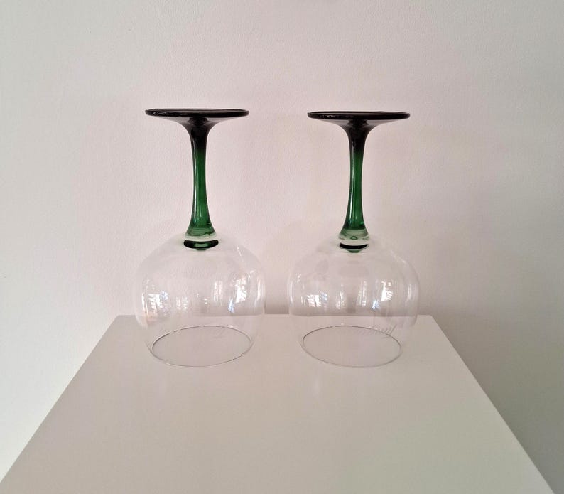 May include: Two clear wine glasses with a green stem and dark rim are displayed upside down. The glasses are on a white surface against a white background. The stem is a vibrant green color, and the rim is a dark, almost black color.