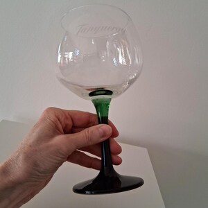 May include: A clear, large-bowled gin glass with a green stem and black base. The word "Tanqueray" is etched on the glass. The glass is held by a person.