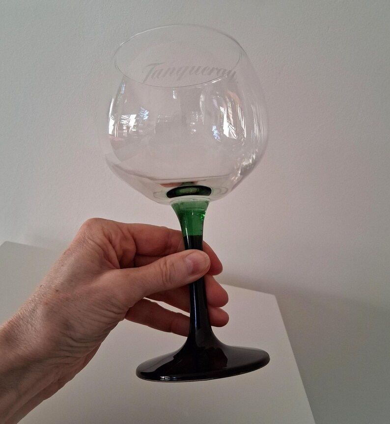 May include: A clear, large-bowled gin glass with a green stem and black base. The word "Tanqueray" is etched on the glass. The glass is held by a person.