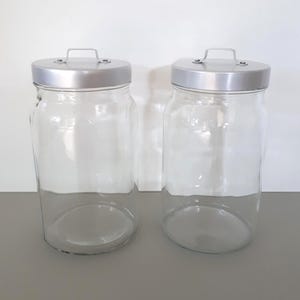 Peut inclure: Deux bocaux de stockage en verre transparent avec des corps cylindriques et des bases plates. Chaque bocal est doté d'un couvercle amovible en métal argenté avec une petite poignée métallique arquée fixée au centre pour faciliter l'ouverture.