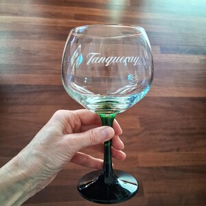 May include: A clear, large gin glass with a green stem and base. The glass has the word "Tanqueray" printed on the front. The glass is held in a hand against a brown wooden background.