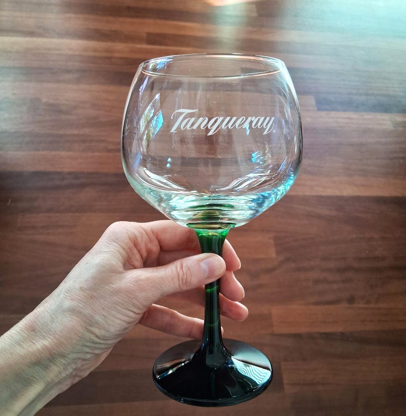 May include: A clear, large gin glass with a green stem and base. The glass has the word "Tanqueray" printed on the front. The glass is held in a hand against a brown wooden background.
