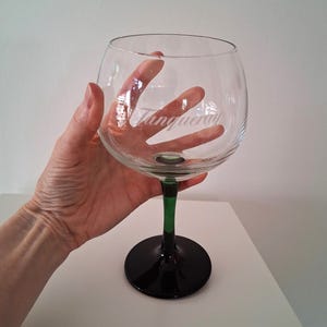 May include: A large, clear glass with a rounded bowl and a black base. The stem is green and black. The word "Tanqueray" is etched on the glass. The glass is designed for cocktails.