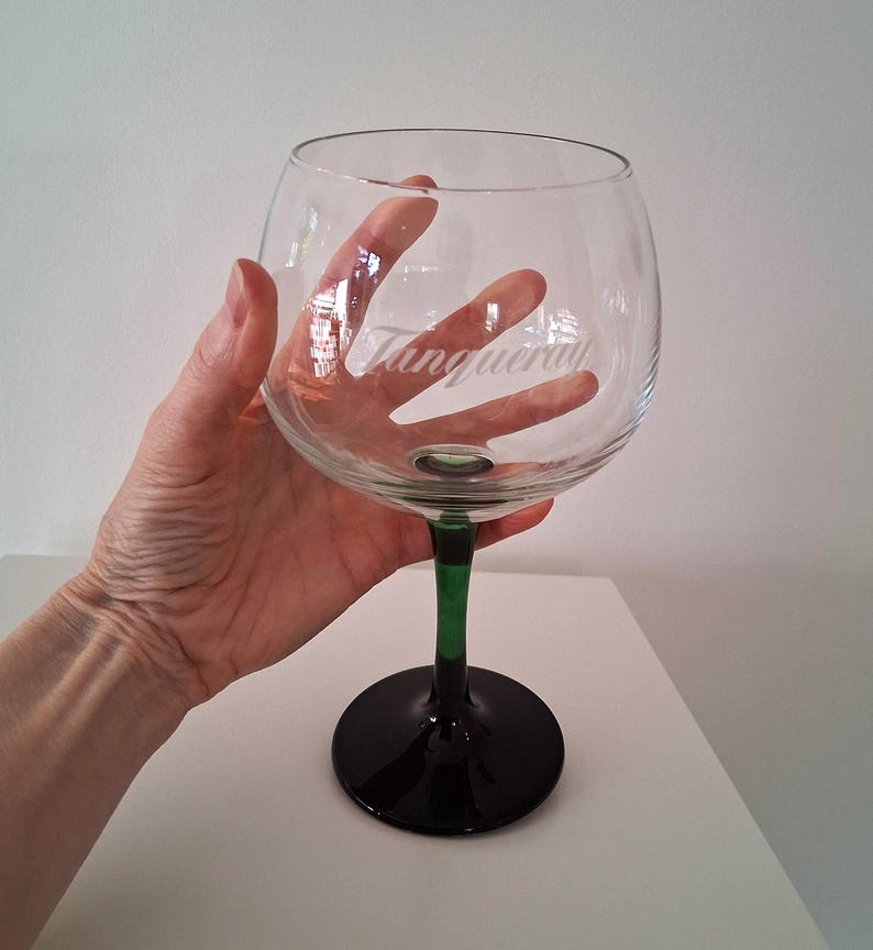May include: A large, clear glass with a rounded bowl and a black base. The stem is green and black. The word "Tanqueray" is etched on the glass. The glass is designed for cocktails.