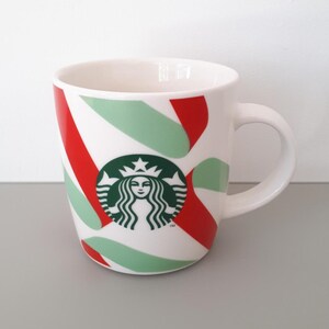 May include: White ceramic mug with a red, green, and white candy cane design. The mug features the Starbucks logo in green on the front. The mug has a curved handle and a glossy finish.