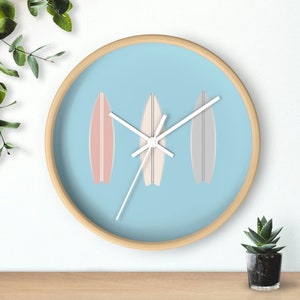 May include: A light blue wall clock with white hands and three surfboard-shaped designs in pink, white, and grey.