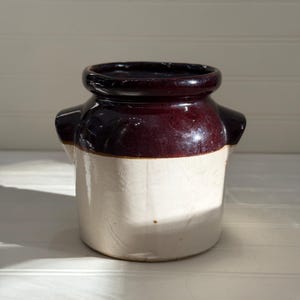 May include: A vintage stoneware crock with a two-tone design. The top portion is a glossy, dark burgundy color, transitioning to a cream-colored base. It features small side handles and a rounded rim. This antique piece is likely used for food storage.